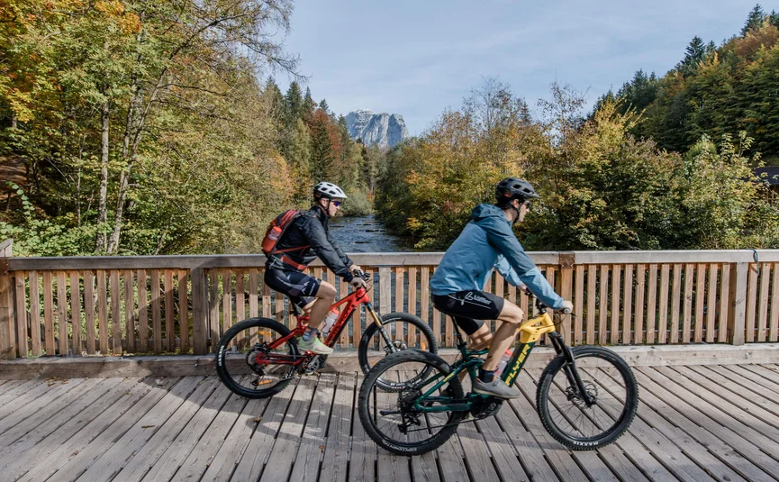 Two cyclists ride on a wooden bridge with forest and river in the background
