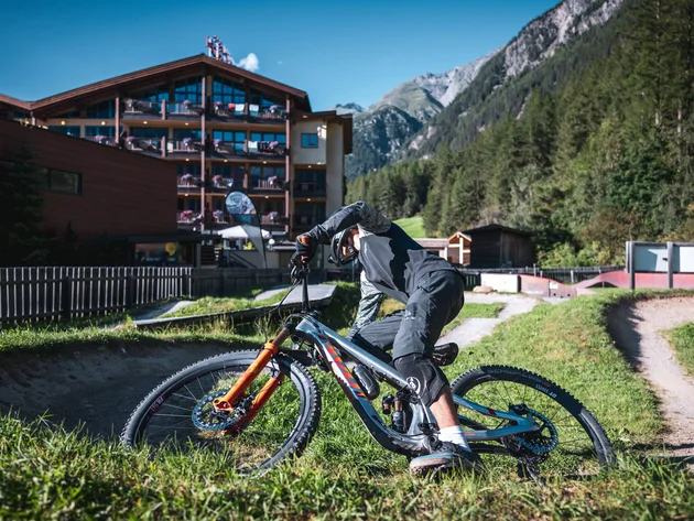 Mountainbiker bei der Abfahrt auf einem Trail vor einem Hotel und Bergen
