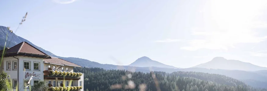 Hotel Maria mit Balkonblumen vor bewaldeten Bergen im Sonnenschein