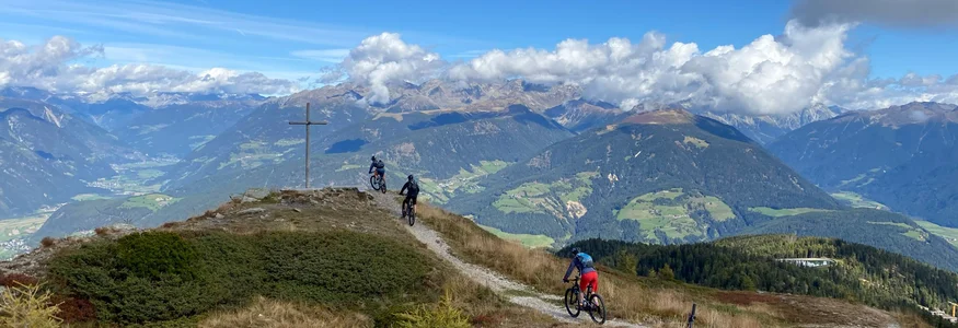Three mountain bikers on a trail with alpine mountains and a cross nearby