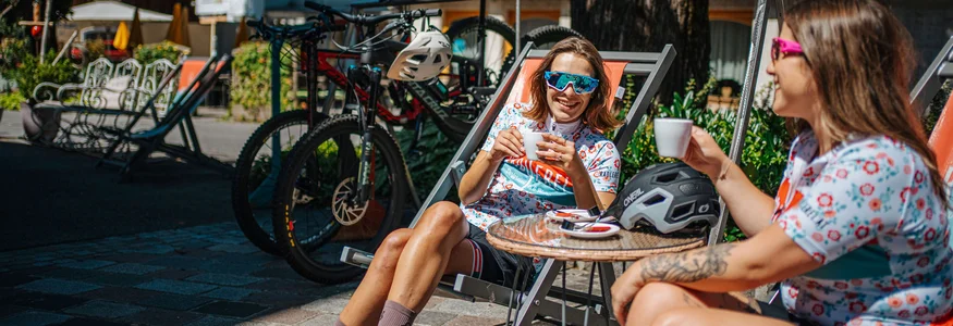 Zwei Frauen in Radbekleidung sitzen draußen mit Kaffee und Fahrrädern im Hintergrund