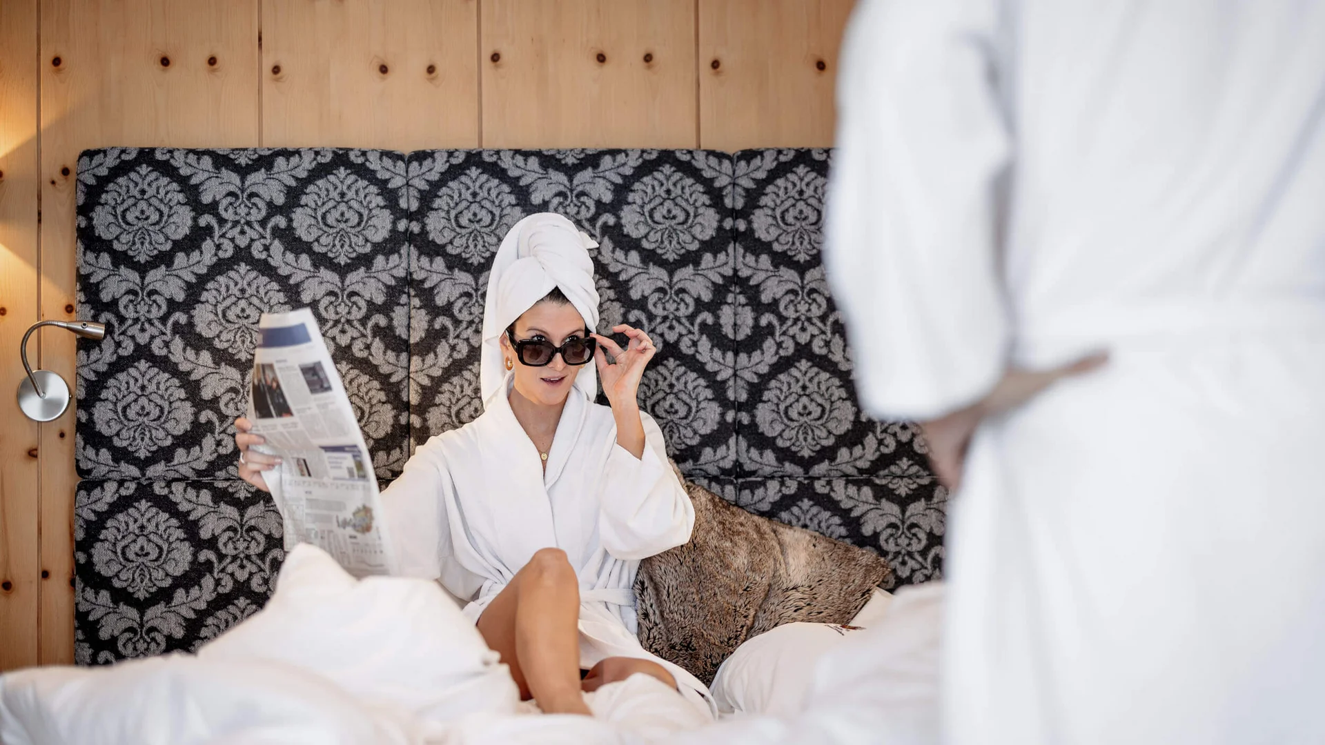 Frau im Bademantel und Handtuch auf dem Kopf liest Zeitung im Bett