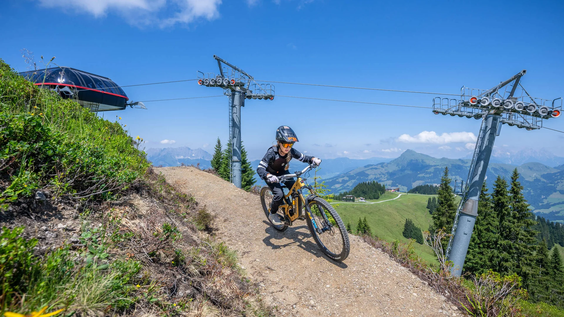 Kind fährt mit Fahrrad auf Bergweg bei Sessellift unter blauem Himmel