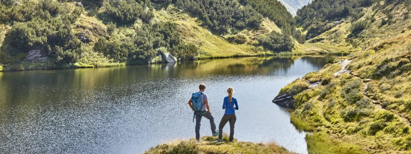 Two hikers standing by a mountain lake in a green alpine landscape