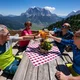 Vier Personen stoßen mit Getränken auf Bergterrasse mit Alpenpanorama an