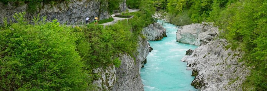 Radfahrer fahren auf einem Weg entlang eines Flusses mit türkisfarbenem Wasser durch eine grüne Schlucht