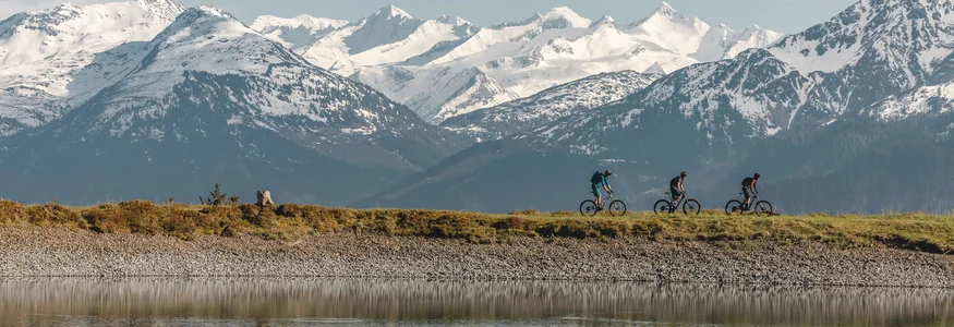 Three mountain bikers biking by a lake with snowy mountains in the background