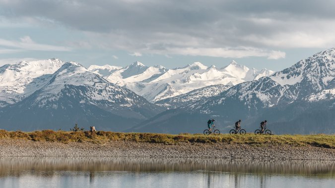 Drei Mountainbiker fahren an einem See vor schneebedeckten Bergen