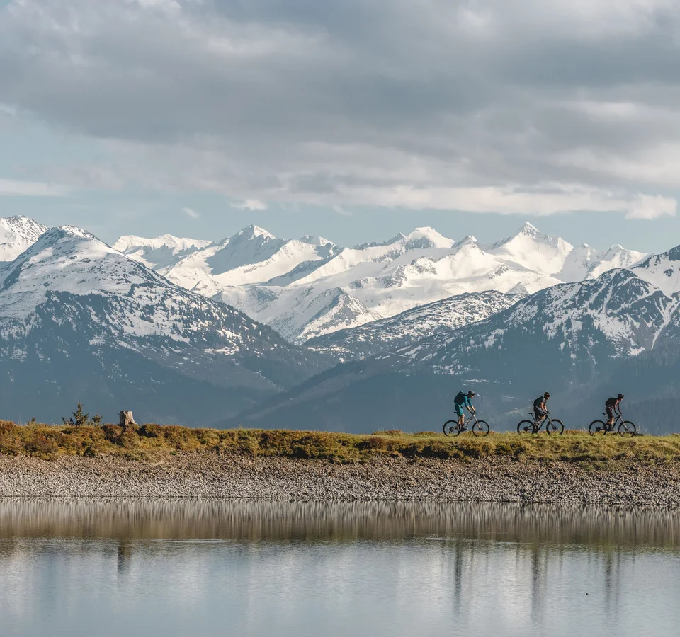 Drei Mountainbiker fahren an einem See vor schneebedeckten Bergen