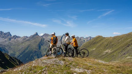 Drei Radfahrer mit Helmen auf einem Berg mit Alpenpanorama