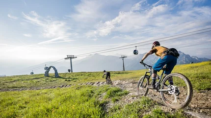 Zwei Mountainbiker fahren auf einem Bergpfad mit Seilbahn und Bergen im Hintergrund