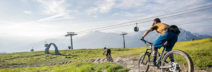 Ein Herbst-Wochenende in San Vigilio © Harald Wisthaler Zwei Mountainbiker fahren auf einem Bergpfad mit Seilbahn und Bergen im Hintergrund
