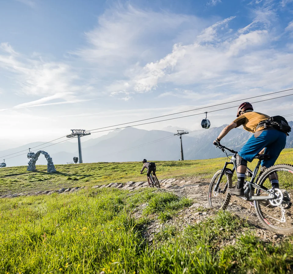 Two mountain bikers riding on a trail with cable cars and mountains in the background