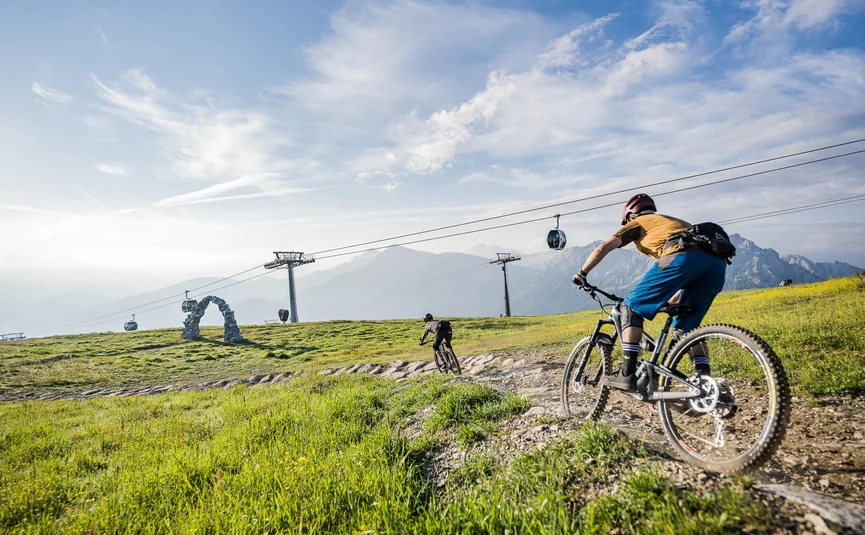 Zwei Mountainbiker fahren auf einem Bergpfad mit Seilbahn und Bergen im Hintergrund
