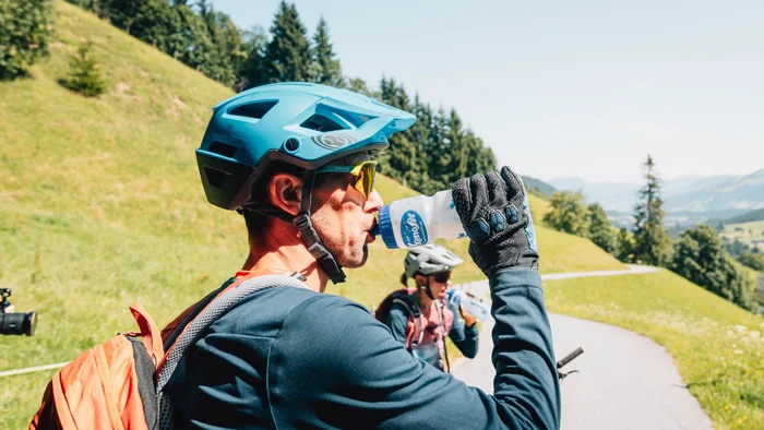Xenofit mineral energy © Patrick Wasshuber Mann mit Fahrradhelm trinkt Wasser auf Bergweg mit grüner Landschaft