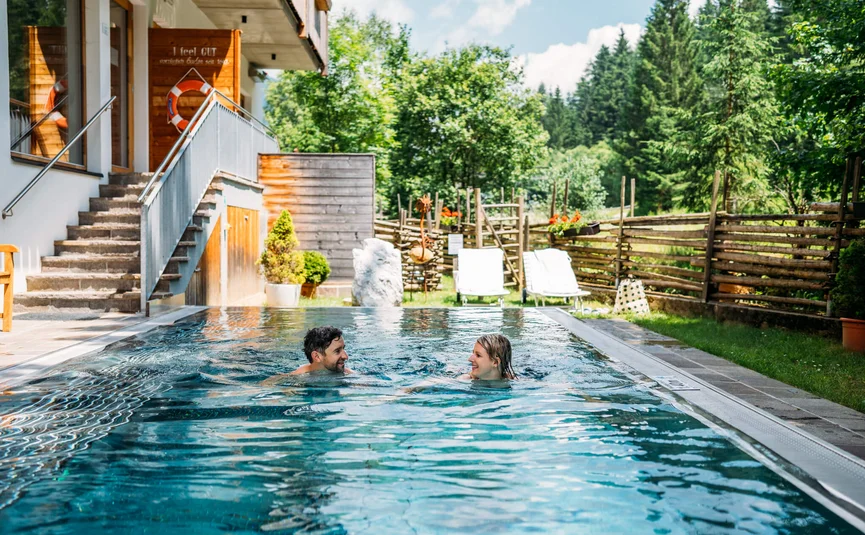 Paar schwimmt im Außenpool neben Holzhaus mit Garten und Bäumen