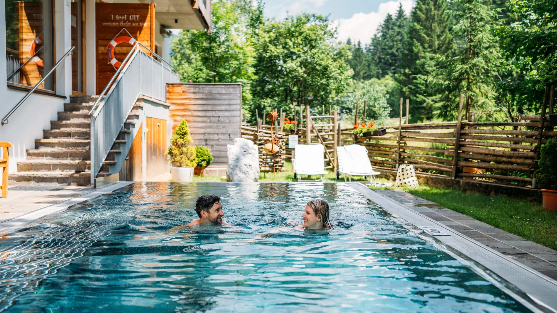 Paar schwimmt im Außenpool neben Holzhaus mit Garten und Bäumen