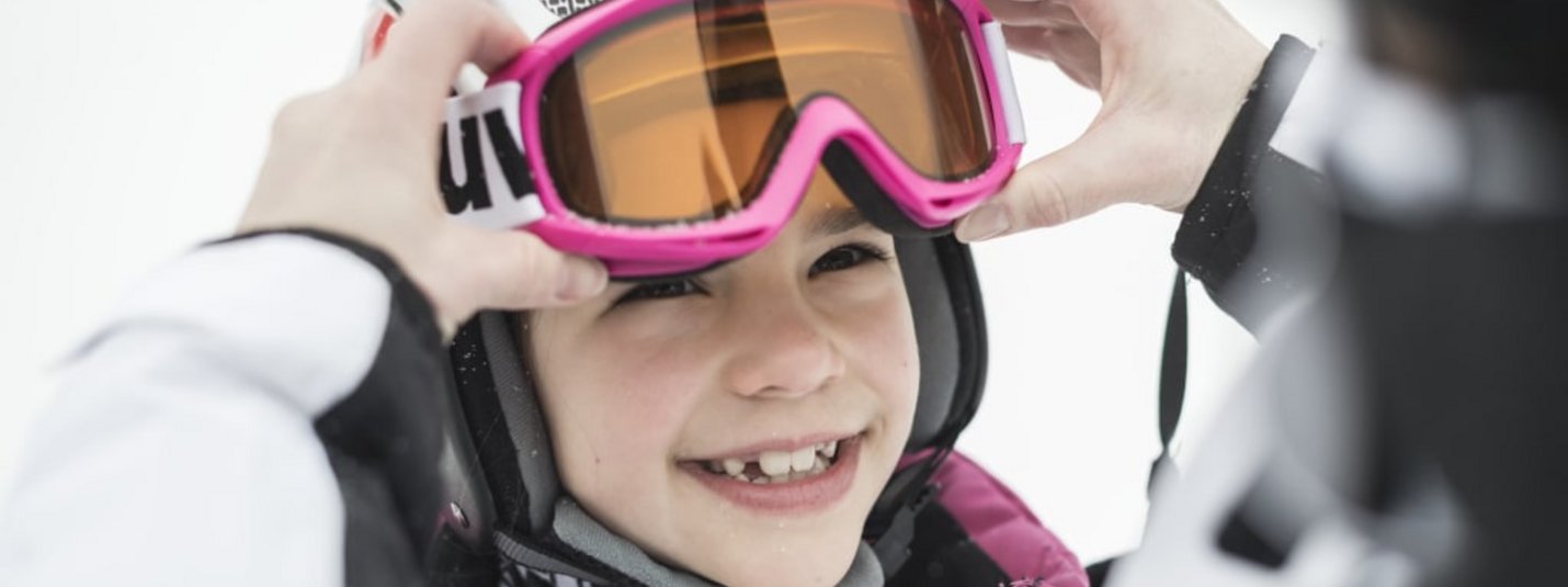Happy Family Skiing Kind mit Skihelm und pinker Skibrille wird beim Anlegen geholfen