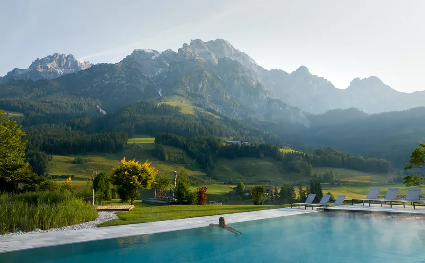 Person entspannt im Pool mit Blick auf bewaldete Berge und grüne Wiesen