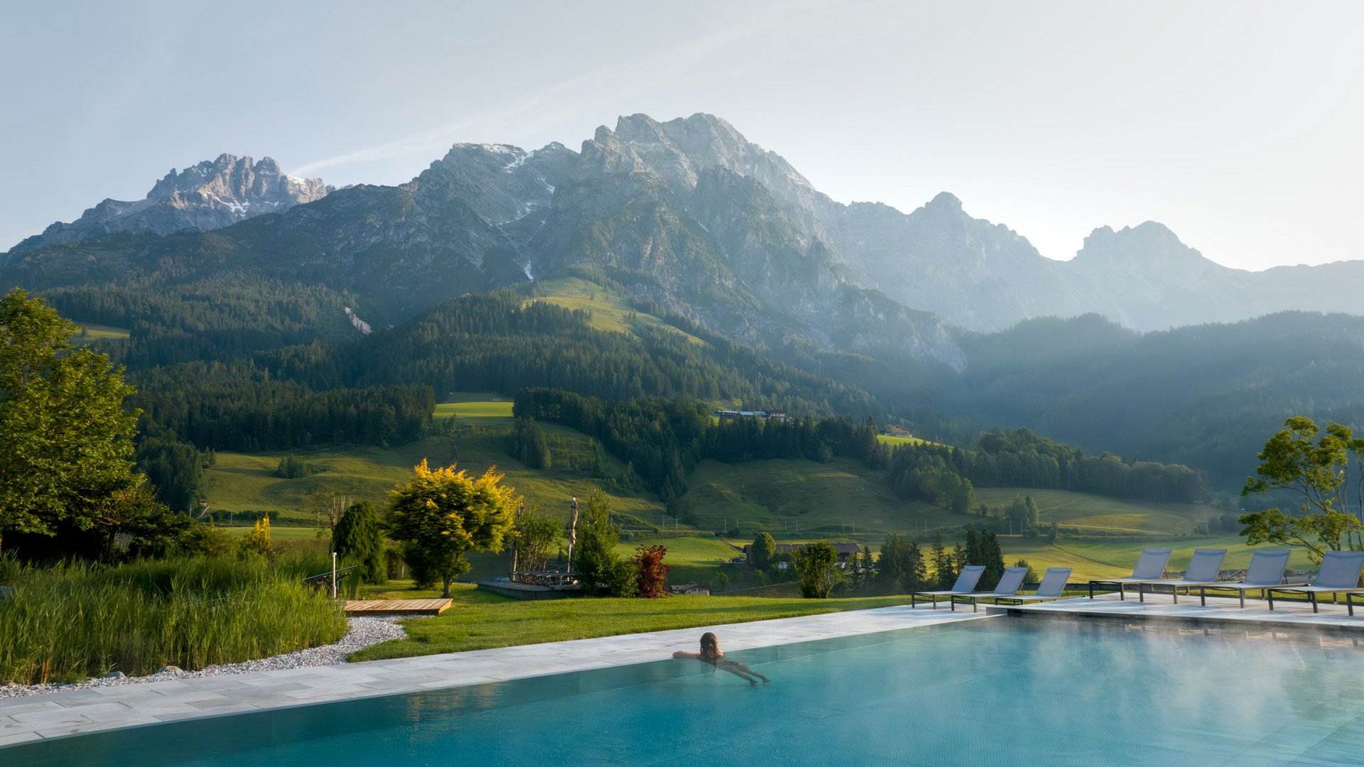 Person entspannt im Pool mit Blick auf bewaldete Berge und grüne Wiesen