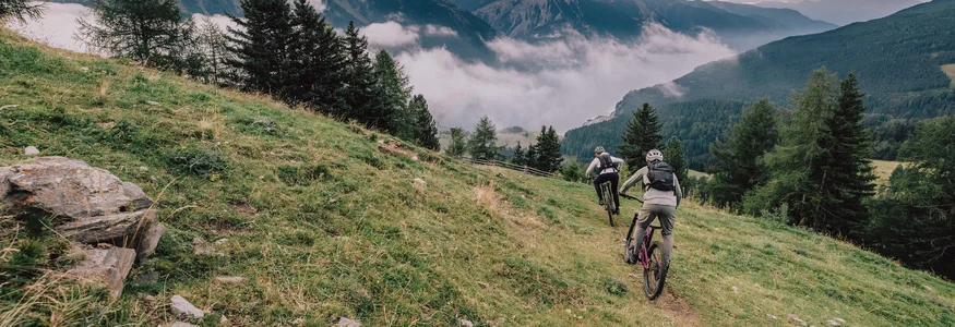 Zwei Mountainbiker auf bergigem Pfad mit Nebel und Bergen im Hintergrund