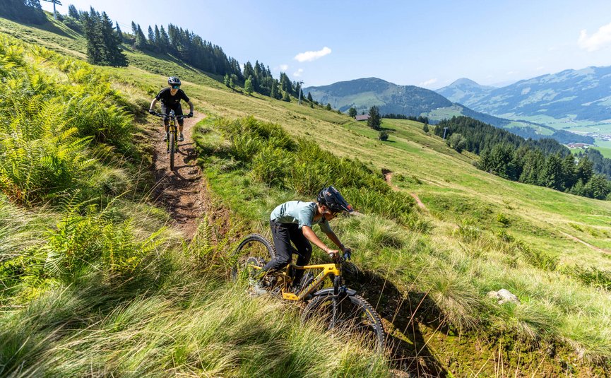 Two mountain bikers riding on a narrow trail in green mountainous landscape