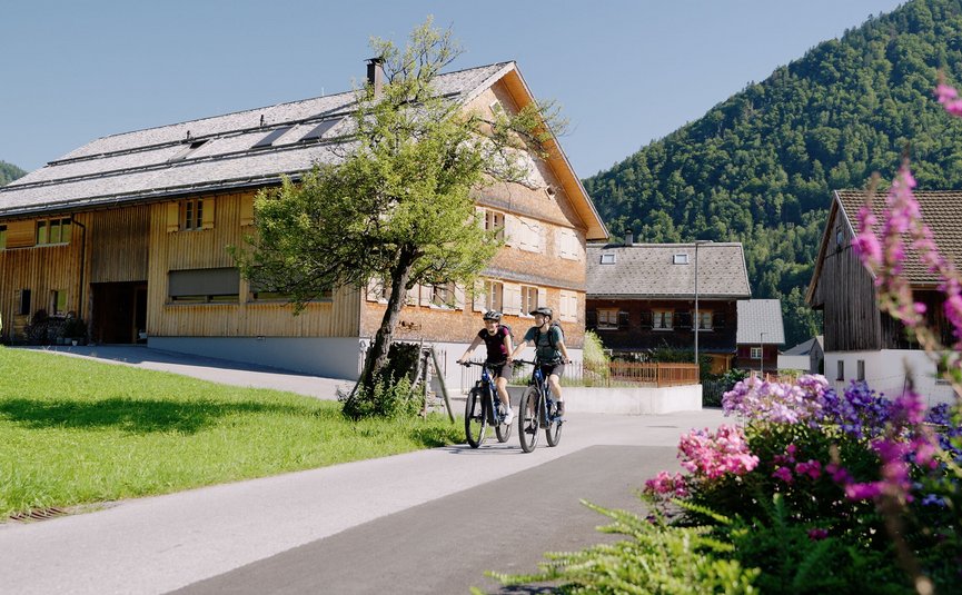 Mountainbikevakantie in het Bregenzerwald © Niklas Kirchler Twee fietsers rijden door een dorp met bergen op een zonnige dag