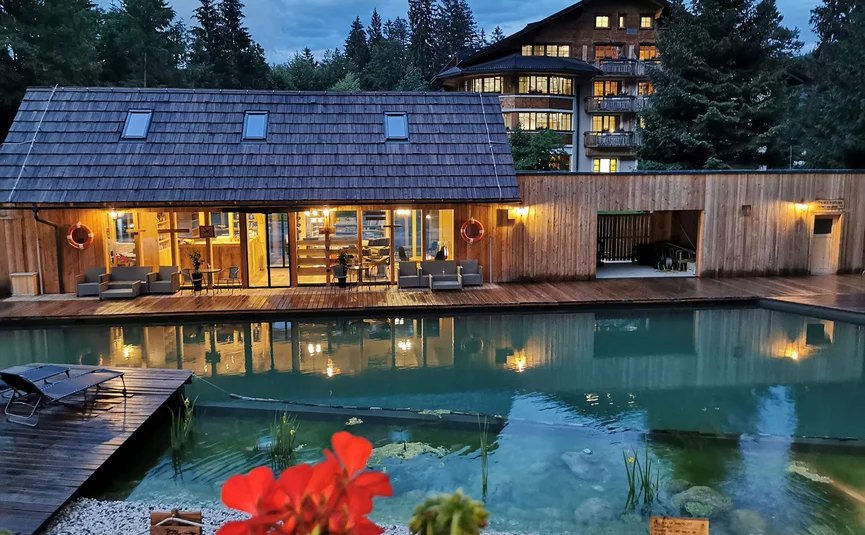 Holzhaus mit beleuchtetem Pool und roten Blumen im Vordergrund bei Abenddämmerung