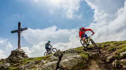 Two mountain bikers riding rocky mountain trail near a wooden cross