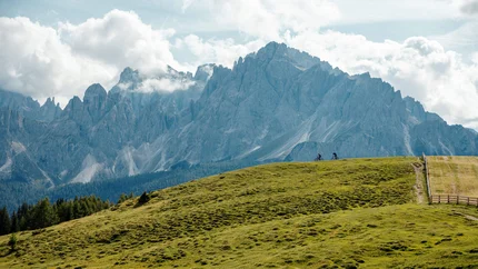 Zwei Radfahrer auf grüner Wiese vor hohen Bergen und wolkigem Himmel