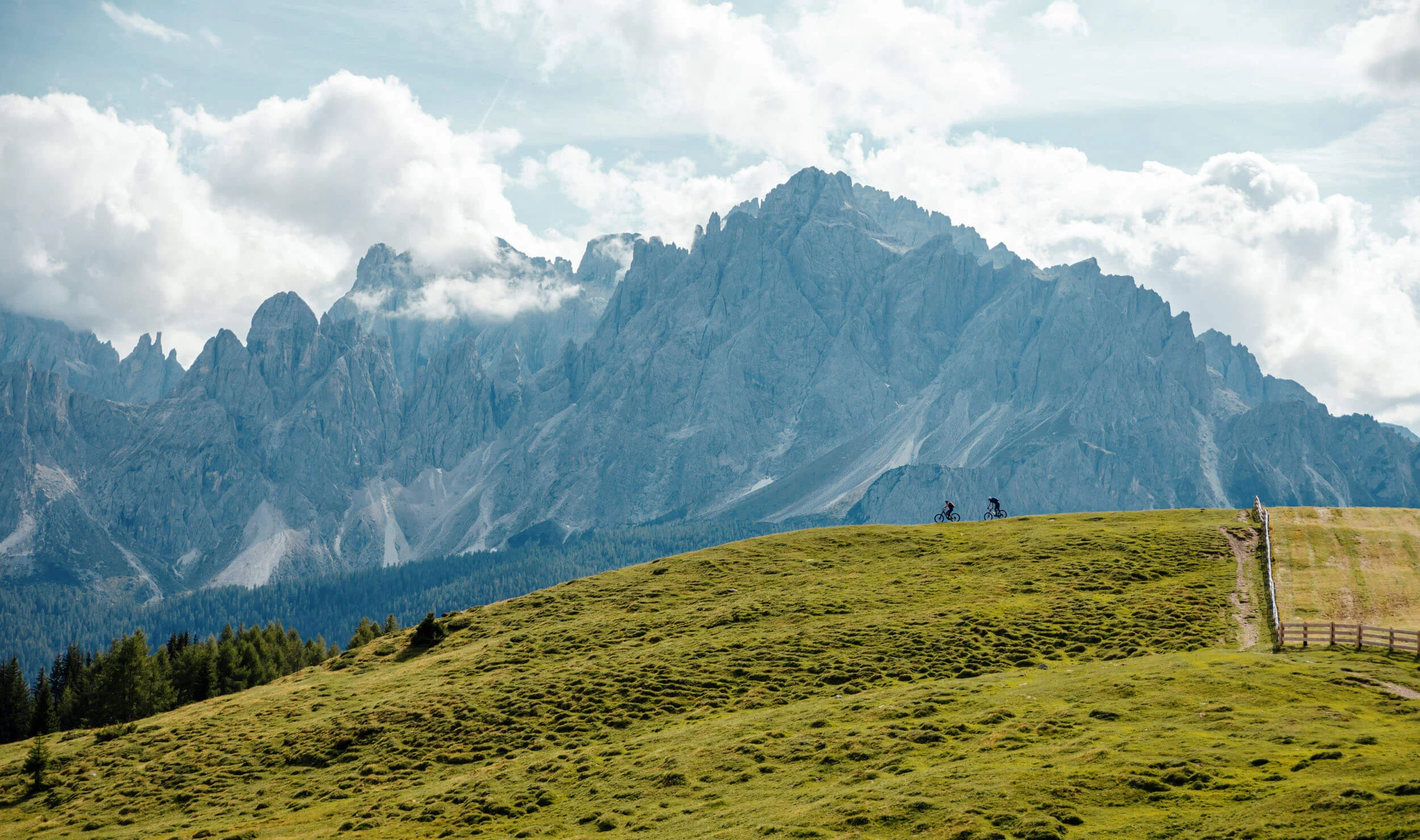 Offers © David Karg Two cyclists on green hill with towering mountains and cloudy sky