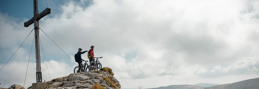 Zwei Mountainbiker auf felsigem Gipfel neben großem Holzkreuz