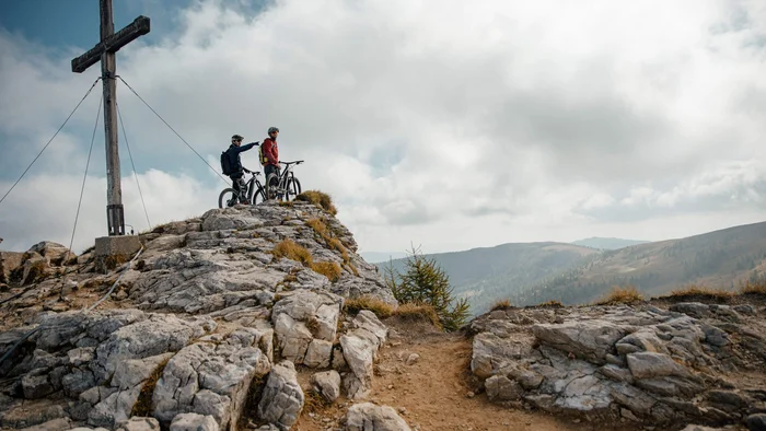 Zwei Mountainbiker auf felsigem Gipfel neben großem Holzkreuz