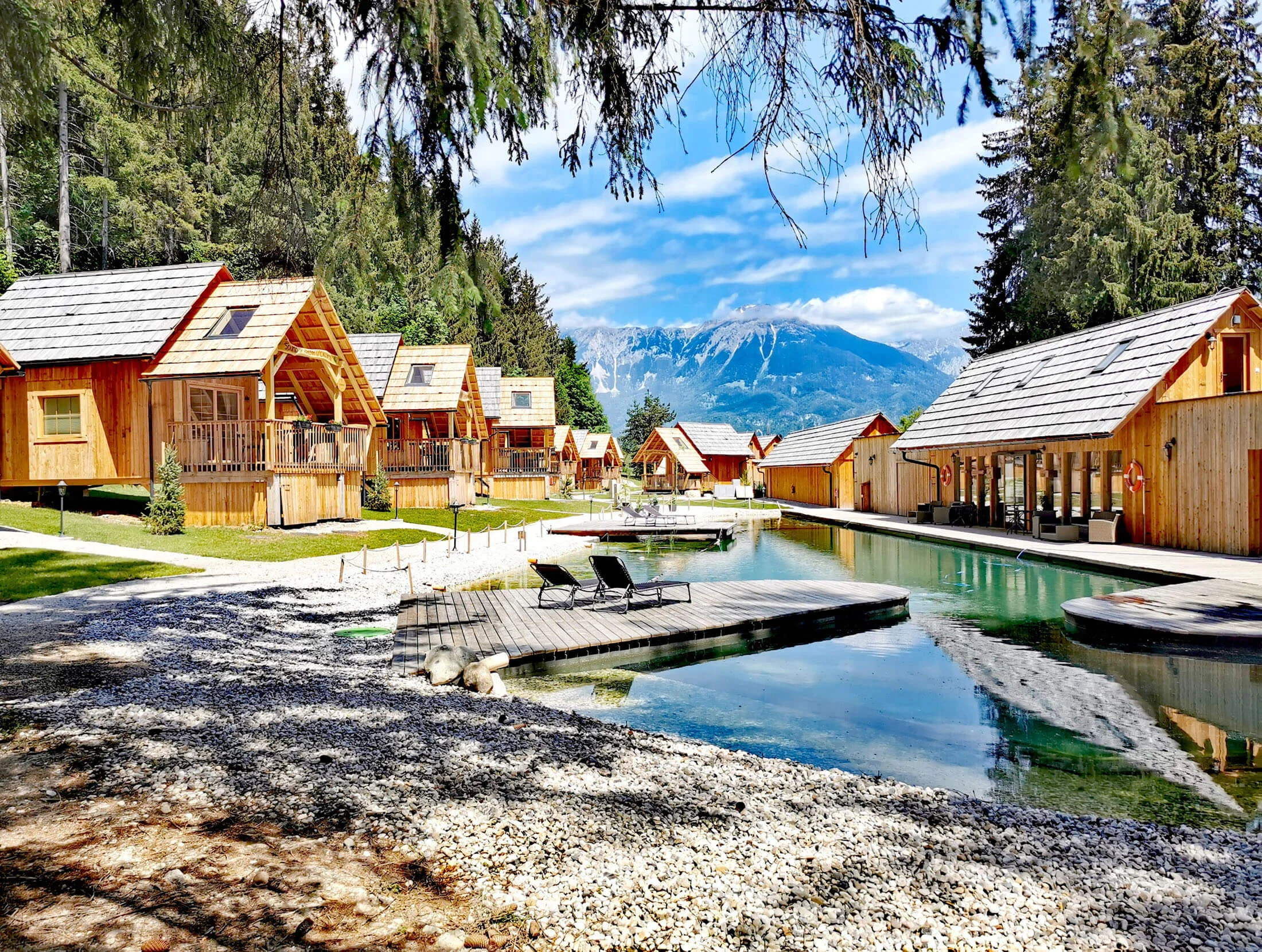 Was steckt hinter dem Umweltengagement von Bikehotels? © Ribno Alpine Resort - Katarina Strgar Holzhütten am See mit Bergblick und zwei Liegestühlen am Steg