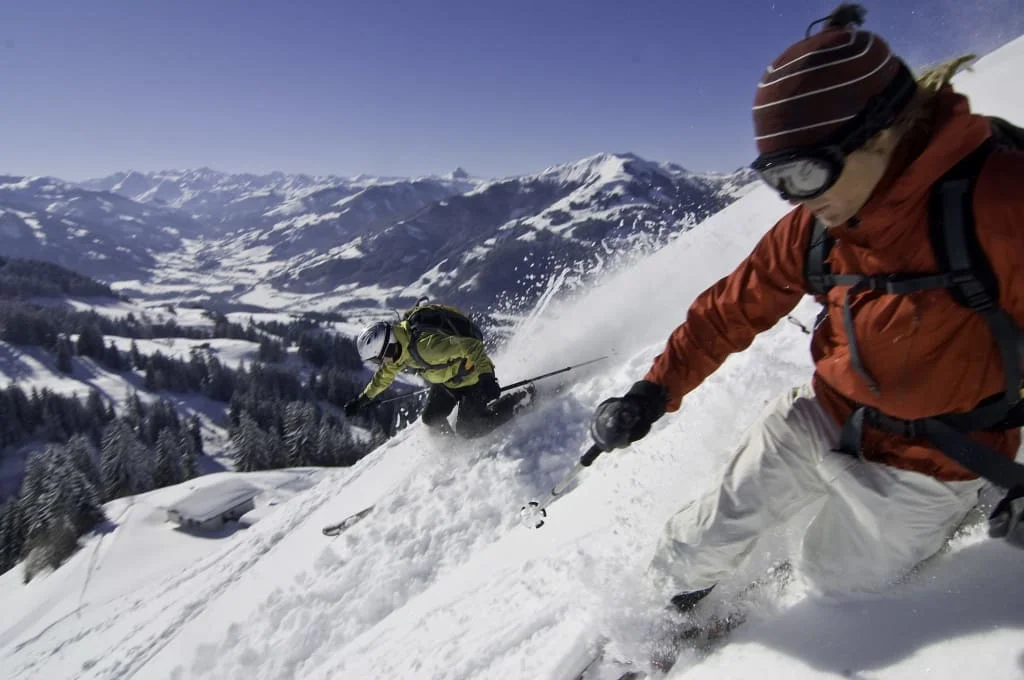 Twee skiërs dalen af door diepe sneeuw met besneeuwde bergen op de achtergrond