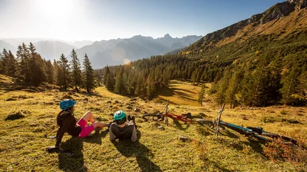 Zwei Radfahrer mit Helmen ruhen auf einer Bergwiese mit Blick auf bewaldete Täler