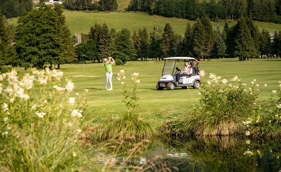 Golfer hitting ball on green golf course with golf cart and trees