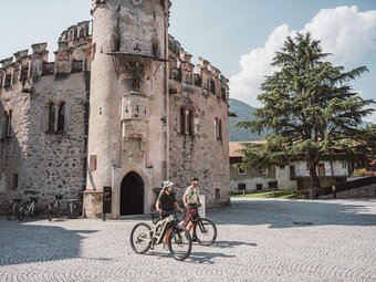 Two cyclists in front of a historic stone tower on a sunny day