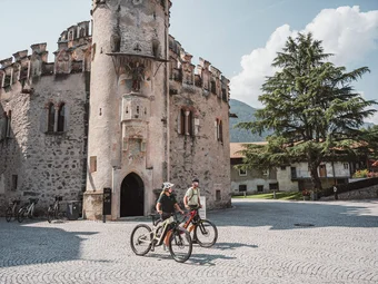 Two cyclists in front of a historic stone tower on a sunny day