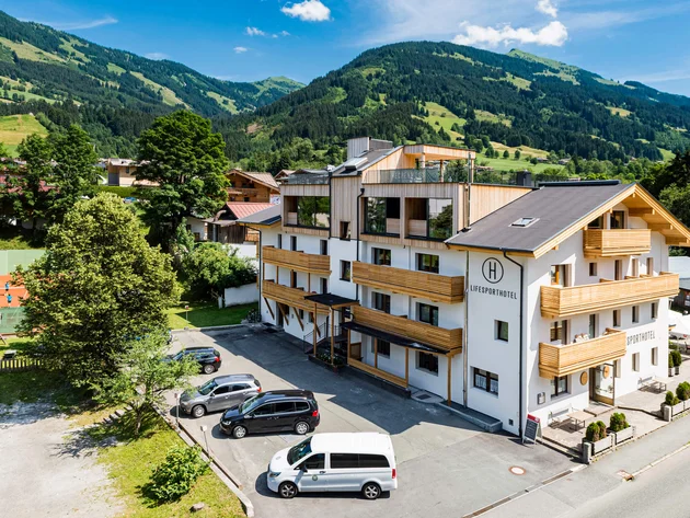 Mit Martina & Flo zum Mountainbiken im LifeSport Hotel Hechenmoos © Multivisual Art Modernes Hotelgebäude mit Bergen im Hintergrund an einem sonnigen Tag