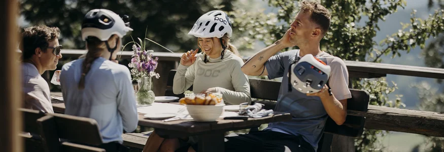 Fahrradfahrer in Helmen sitzen draußen an einem Holztisch und lachen