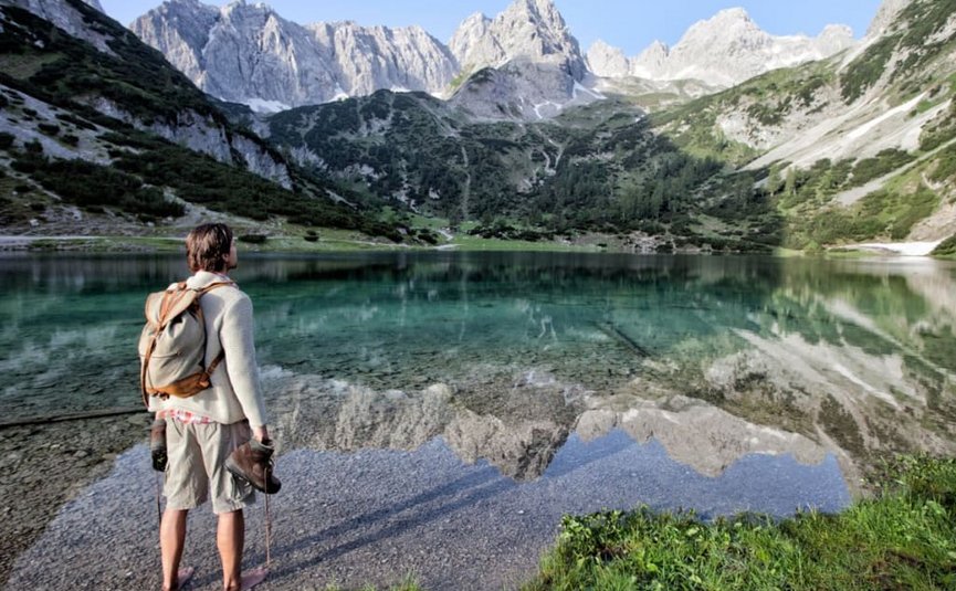 Hiker stands barefoot at mountain lake with clear water reflection and peaks