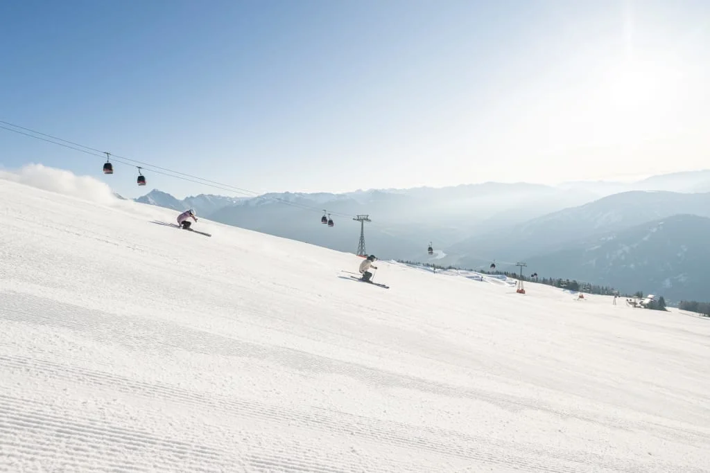 Skiërs op een besneeuwde piste met een kabelbaan en bergen op de achtergrond