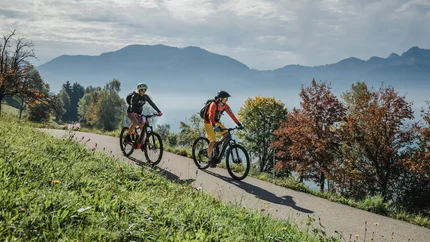 Zwei Radfahrer auf einem Weg in bergiger Herbstlandschaft mit Seen und Bäumen