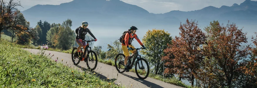 Zwei Radfahrer auf einem Weg in bergiger Herbstlandschaft mit Seen und Bäumen