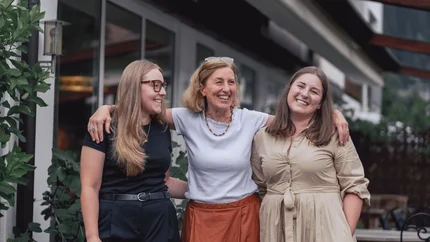 Drie lachende vrouwen staan arm in arm voor een gebouw