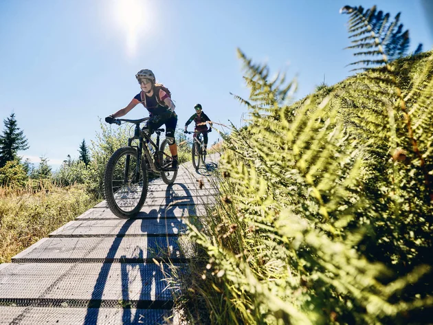 Happy Birthday EPIC Bike Park! © Tobias Köhler Zwei Mountainbiker fahren auf einem Holzsteg durch eine sonnige Landschaft