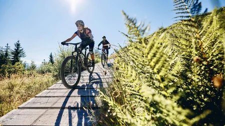 Happy Birthday EPIC Bike Park! © Tobias Köhler Zwei Mountainbiker fahren auf einem Holzsteg durch eine sonnige Landschaft