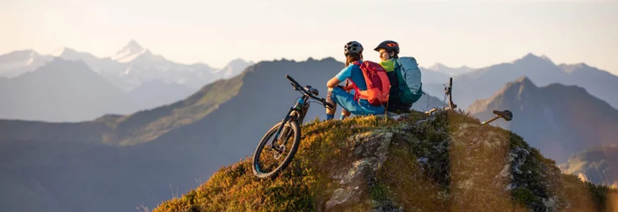 Zwei Radfahrer mit Rucksäcken sitzen auf einem Berggipfel mit Blick auf Berge