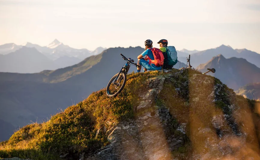 Two cyclists with backpacks sitting on a mountain peak overlooking mountains